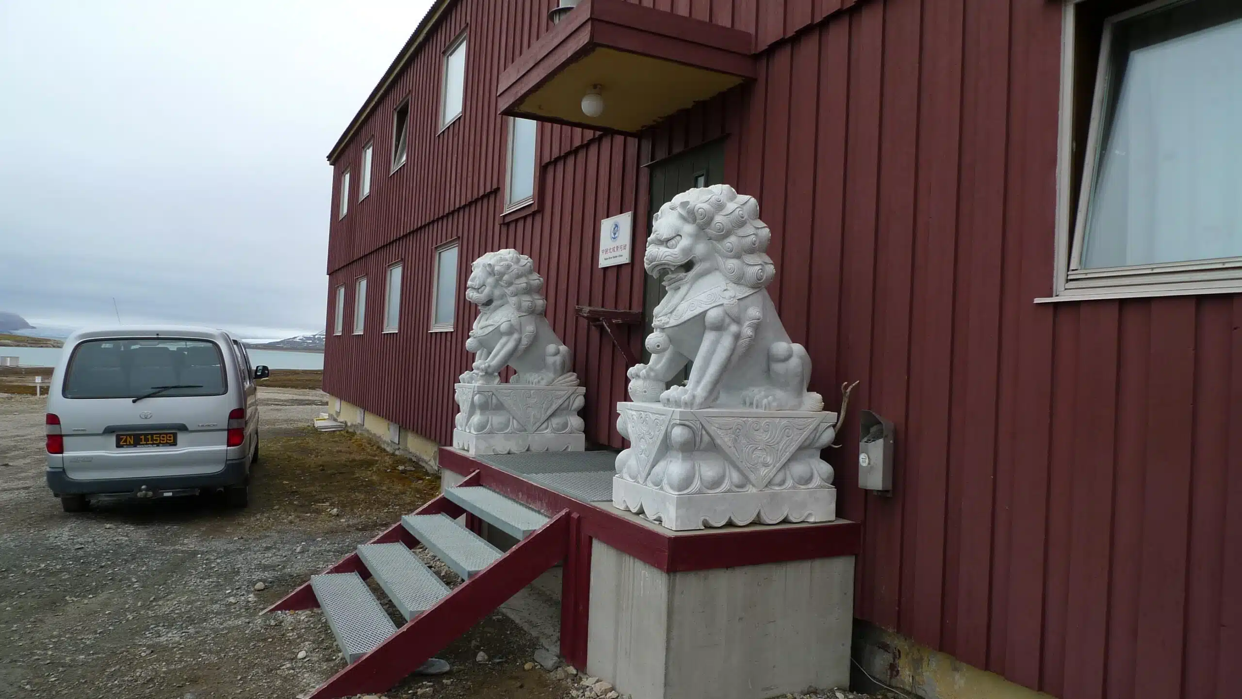 Two white Chinese dragon statues in front of a red building, a gray van parked outside, gray Arctic weather