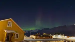 A yellow building against a dark blue starry sky with green northern lights and other cabins and snow in the background