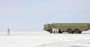 Soldiers in white uniforms stand on snowy ground in front of a green truck