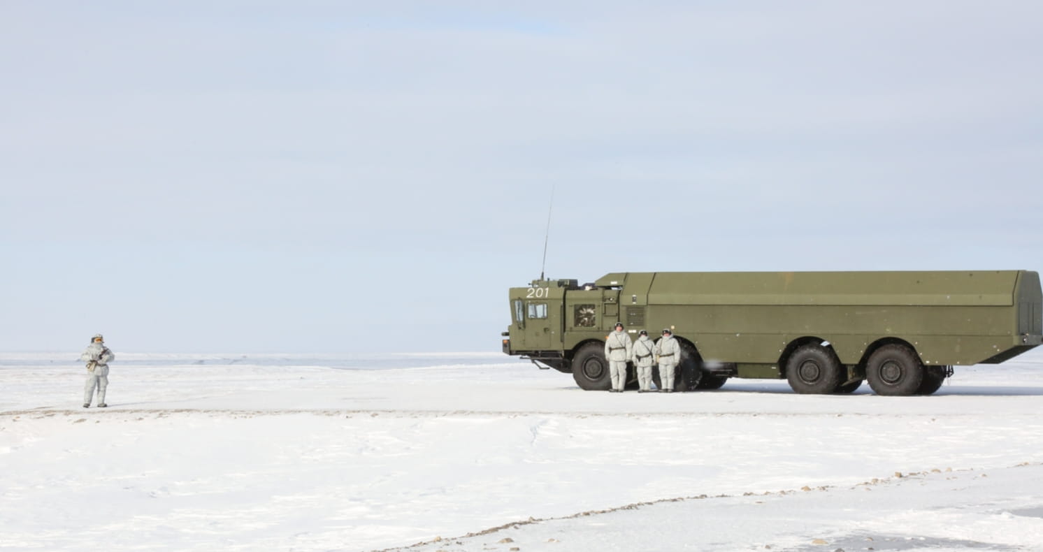 Soldiers in white uniforms stand on snowy ground in front of a green truck