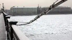 Snow-covered view of the Neva River in St. Petersburg, Russia, with historic buildings and a bridge with snow on the railing