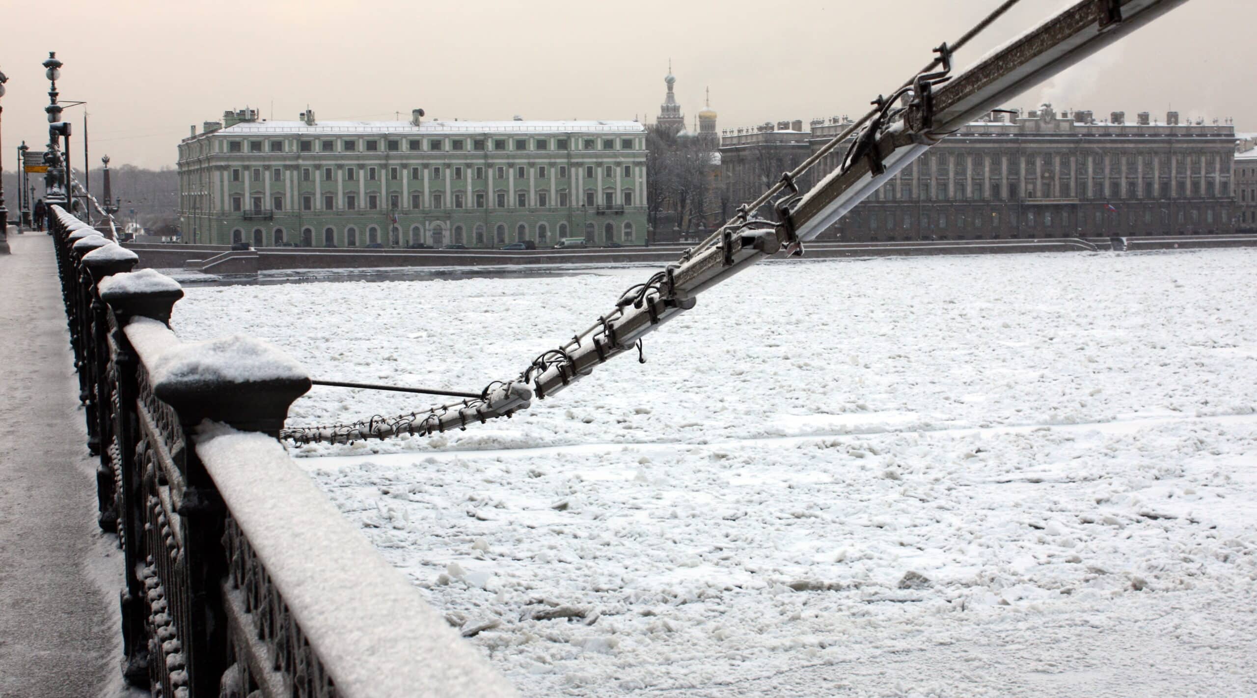 Snow-covered view of the Neva River in St. Petersburg, Russia, with historic buildings and a bridge with snow on the railing