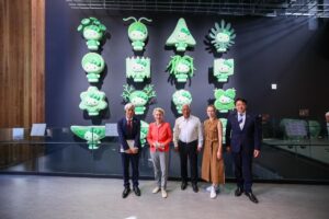 Von der Leyen, da Costa, Kallas and Ishiba standing in a modern indoor exhibition space, posing in front of a wall display featuring multiple green cartoon-style characters arranged in rows