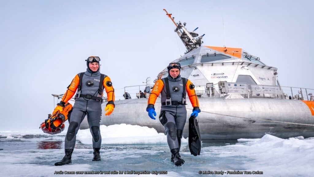 Two crew members in cold-water diving suits walk across Arctic sea ice in front of the Tara Ocean research vessel during a hull inspection expedition