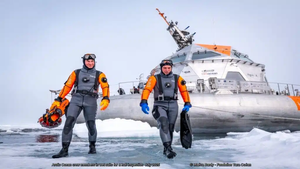 Two crew members in cold-water diving suits walk across Arctic sea ice in front of the Tara Ocean research vessel during a hull inspection expedition