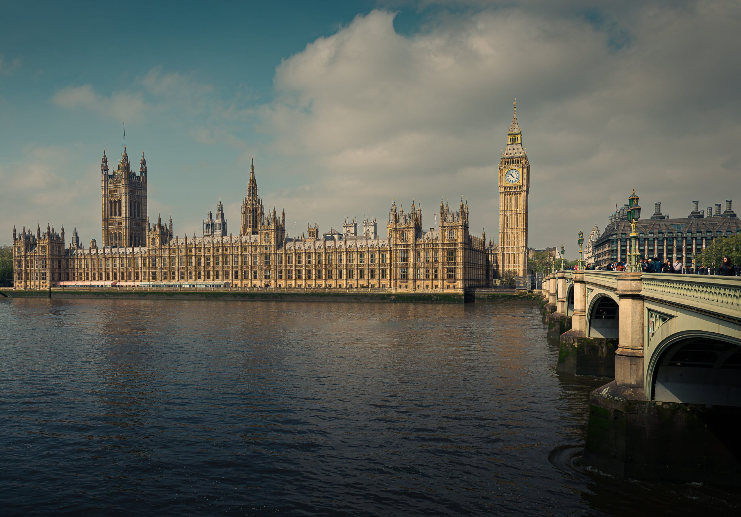 A wide view of the Palace of Westminster and the clock tower known as Big Ben beside the River Thames in London. The historic Gothic-style buildings are lit by soft daylight under a partly cloudy sky, with Westminster Bridge extending into the foreground on the right