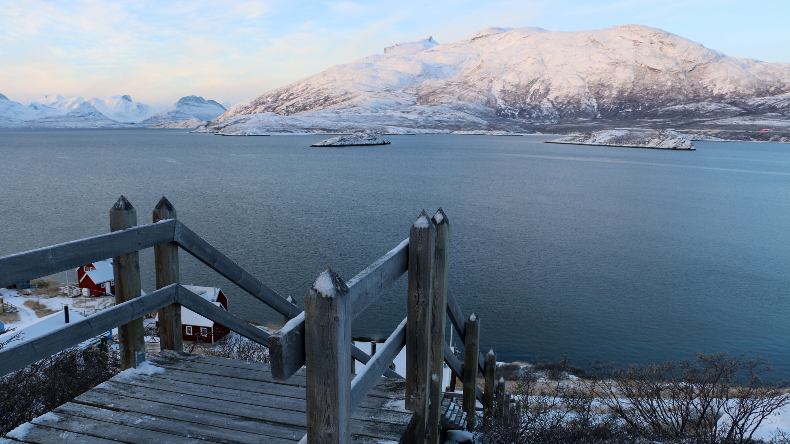 A snowy mountain landscape rises across a calm fjord, viewed from a wooden staircase overlooking red houses along the winter shoreline