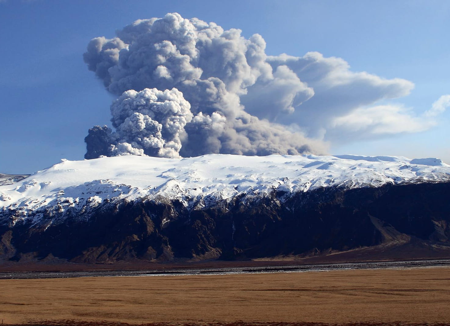 icture shows a cloud of ash rising from the Eyjafjallajökull eruption of Iceland in the background in 2010 during a sunny day in the south of Iceland. In the middle ground, there are small mountains covered by ice and snow that Eyjafjallajökull is erupting from. In the foreground, there is a river and a tan-colored grass field