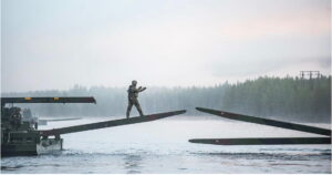 A soldier in combat gear walks across a partially assembled pontoon bridge over a misty Norwegian river during Exercise Trident Juncture 2018, a large-scale NATO interoperability exercise conducted in Norway
