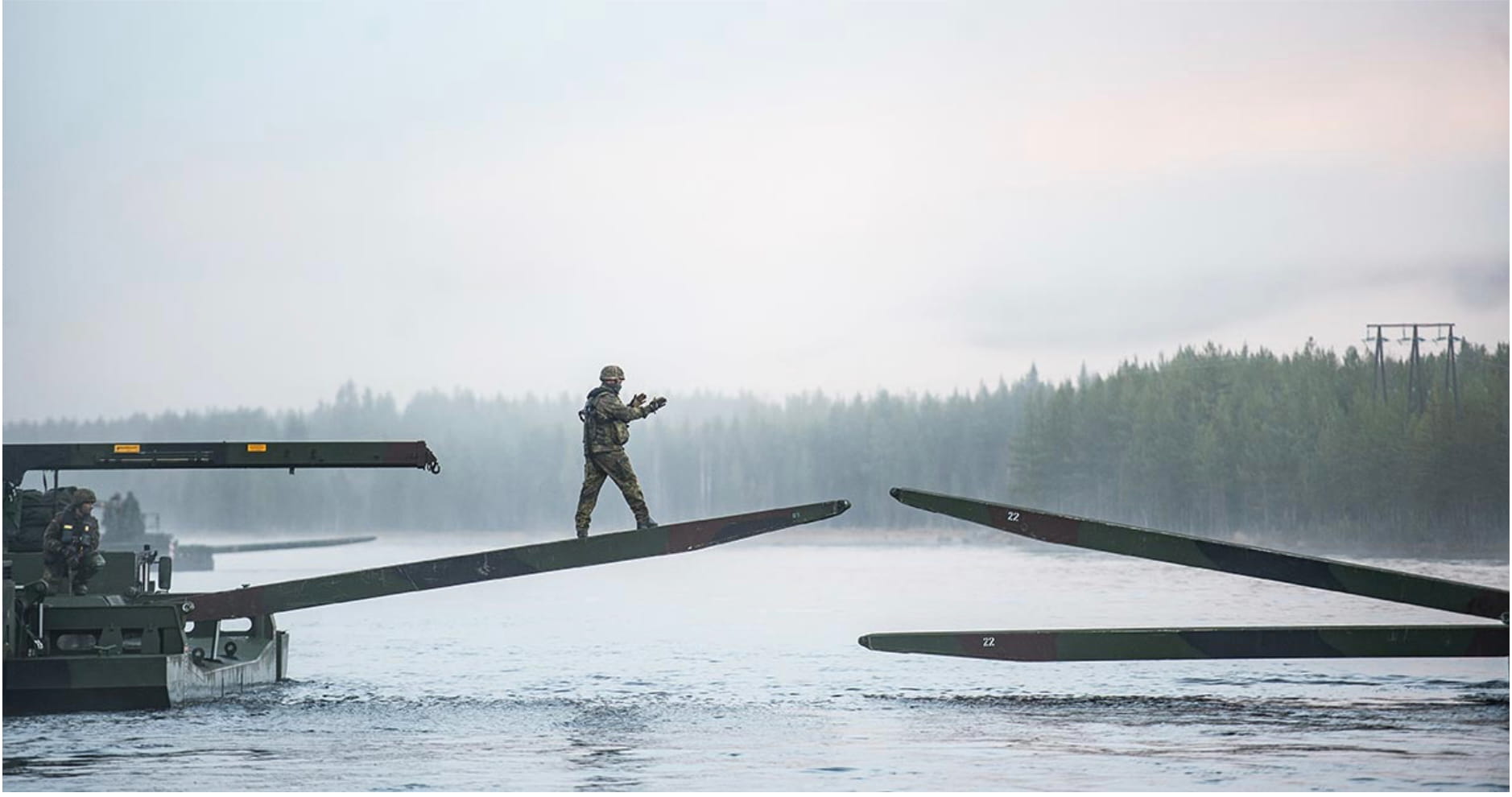 A soldier in combat gear walks across a partially assembled pontoon bridge over a misty Norwegian river during Exercise Trident Juncture 2018, a large-scale NATO interoperability exercise conducted in Norway