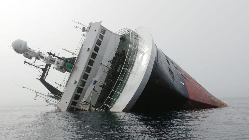 A large ship is heavily listing to its starboard side in calm water, with much of the hull partially submerged. The white superstructure leans at a steep angle, while the black-and-red hull disappears beneath the surface. The overcast sky and still sea emphasize the vessel's distress, and no people or rescue craft are clearly visible