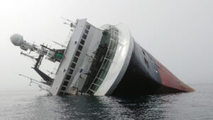 A large ship is heavily listing to its starboard side in calm water, with much of the hull partially submerged. The white superstructure leans at a steep angle, while the black-and-red hull disappears beneath the surface. The overcast sky and still sea emphasize the vessel's distress, and no people or rescue craft are clearly visible