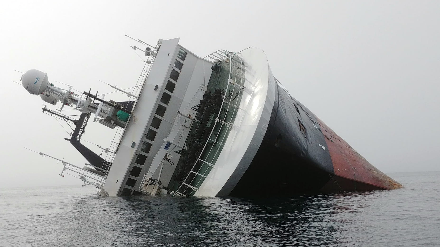 A large ship is heavily listing to its starboard side in calm water, with much of the hull partially submerged. The white superstructure leans at a steep angle, while the black-and-red hull disappears beneath the surface. The overcast sky and still sea emphasize the vessel's distress, and no people or rescue craft are clearly visible