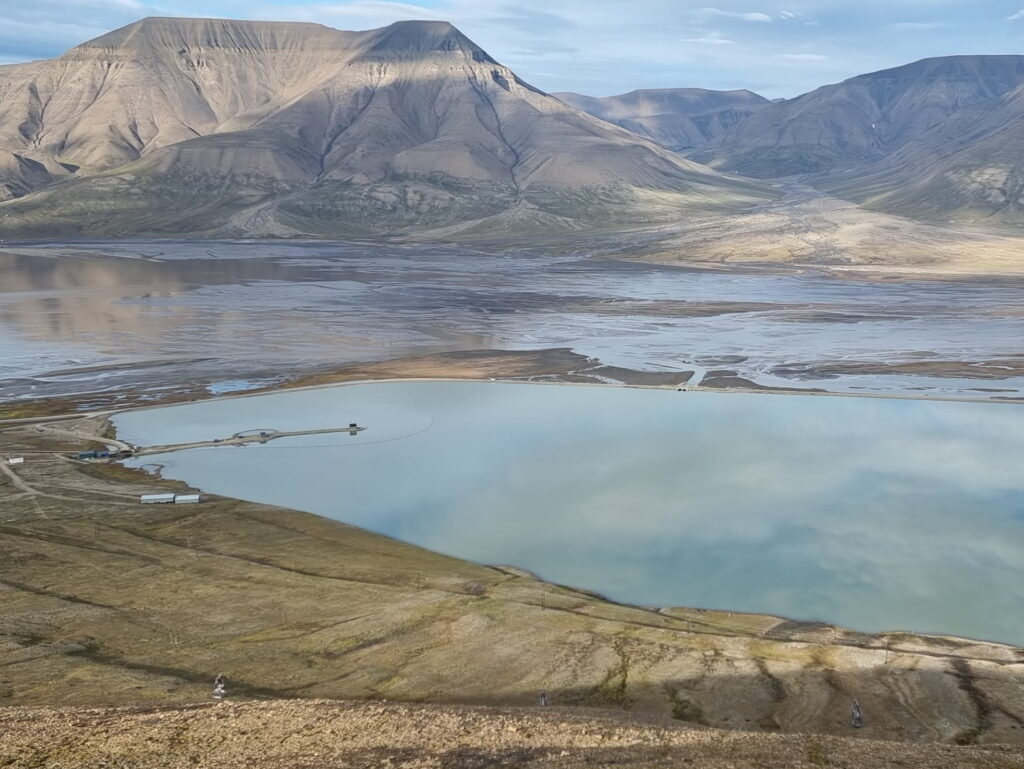 A wide view of a tranquil Arctic landscape featuring a pale blue lake in the foreground, bordered by tundra and a narrow causeway extending into the water. Beyond the lake lies a broad river delta with braided channels stretching across a flat valley. Steep, gray, flat-topped mountains dominate the background, their slopes highlighted by patches of sunlight beneath a partly cloudy sky. The scene is expansive, remote, and largely treeless, with no visible buildings except a few small structures near the water's edge