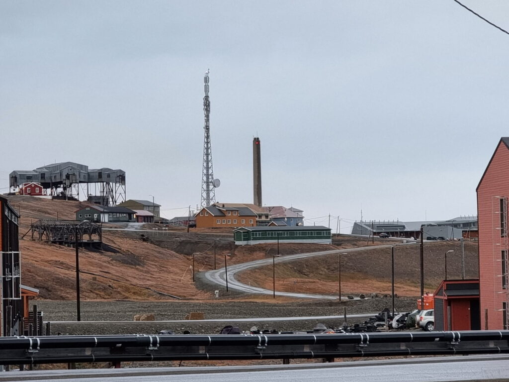 A winding road leads through a small Arctic town with colorful buildings under an overcast sky. A tall communications tower and a smokestack rise above the settlement, while industrial structures sit on a nearby hillside with mostly snow-free, brown terrain