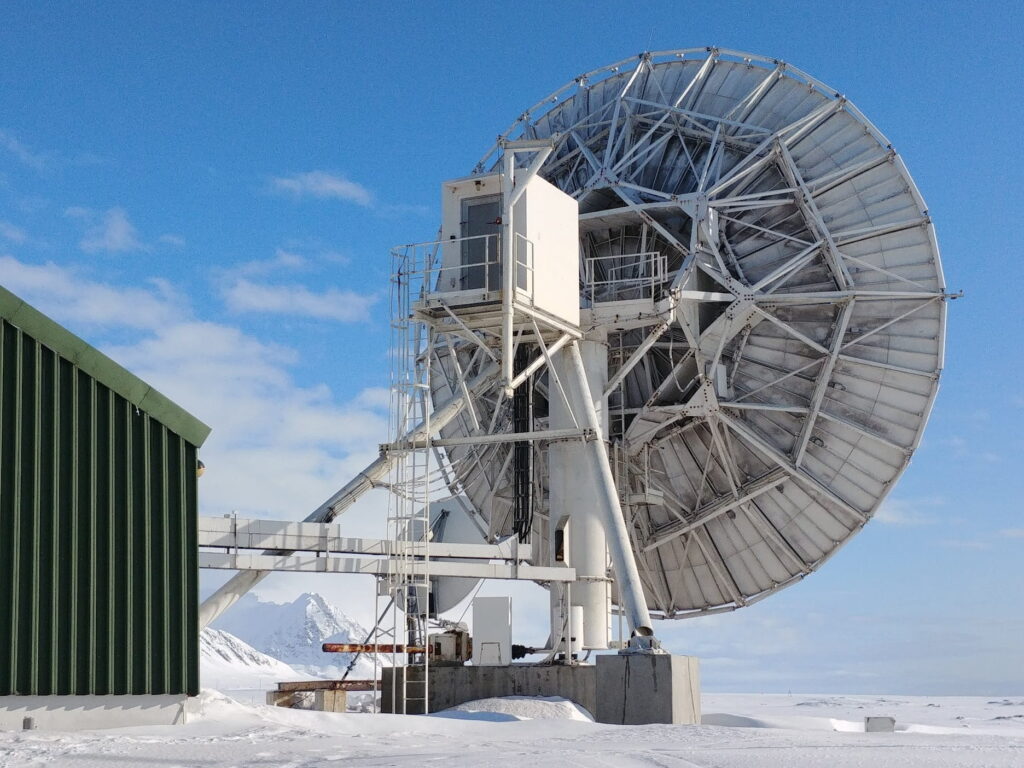 A large white parabolic satellite dish stands on a steel support structure in a snow-covered Arctic landscape. A green industrial building sits to the left, while snow-covered mountains rise in the distance under a bright blue sky with scattered clouds. The antenna dominates the scene, emphasizing the remote research or communications facility