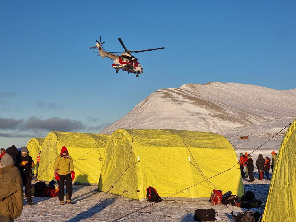 A red-and-white rescue helicopter hovers above a snowy field with bright yellow expedition tents below. Several people in winter clothing stand around the camp, while snow-covered mountains rise in the background under a clear blue sky lit by low-angle sunlight