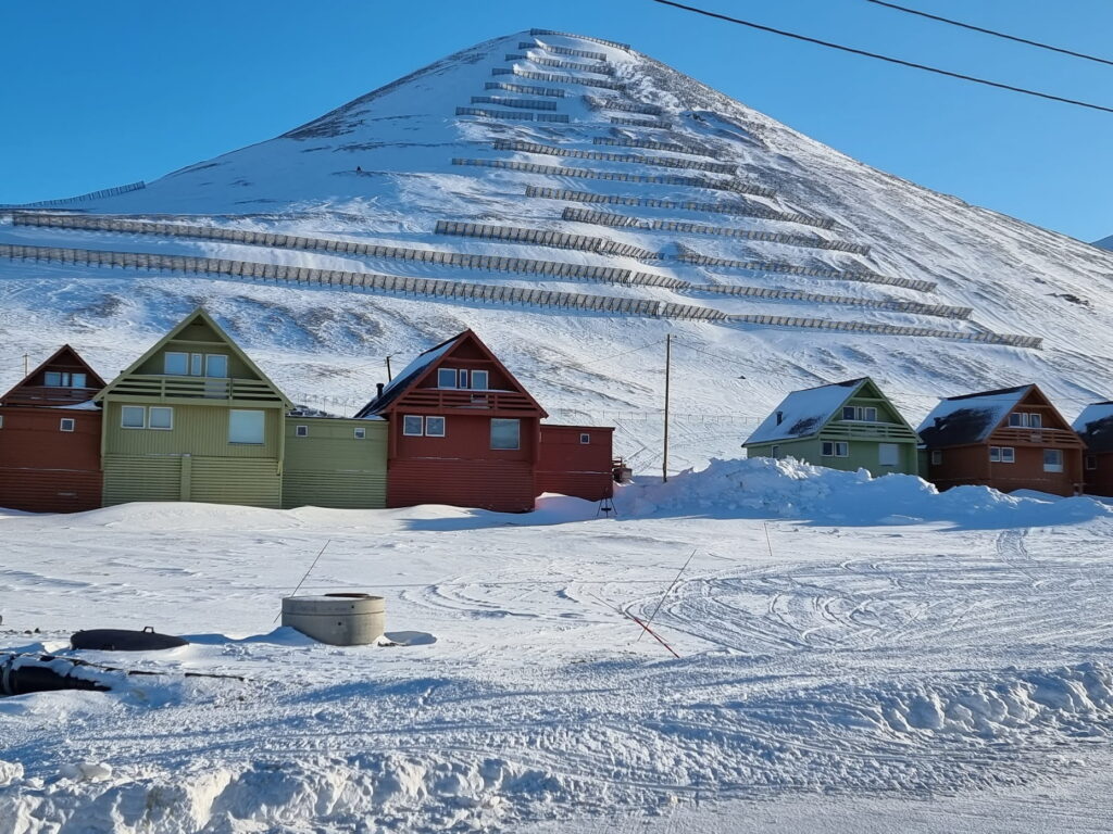 A row of colorful wooden houses sits in a snow-covered settlement at the base of a steep, snow-covered mountain. Multiple avalanche barriers line the mountainside in horizontal rows, while snowbanks and vehicle tracks cover the foreground under a clear blue sky