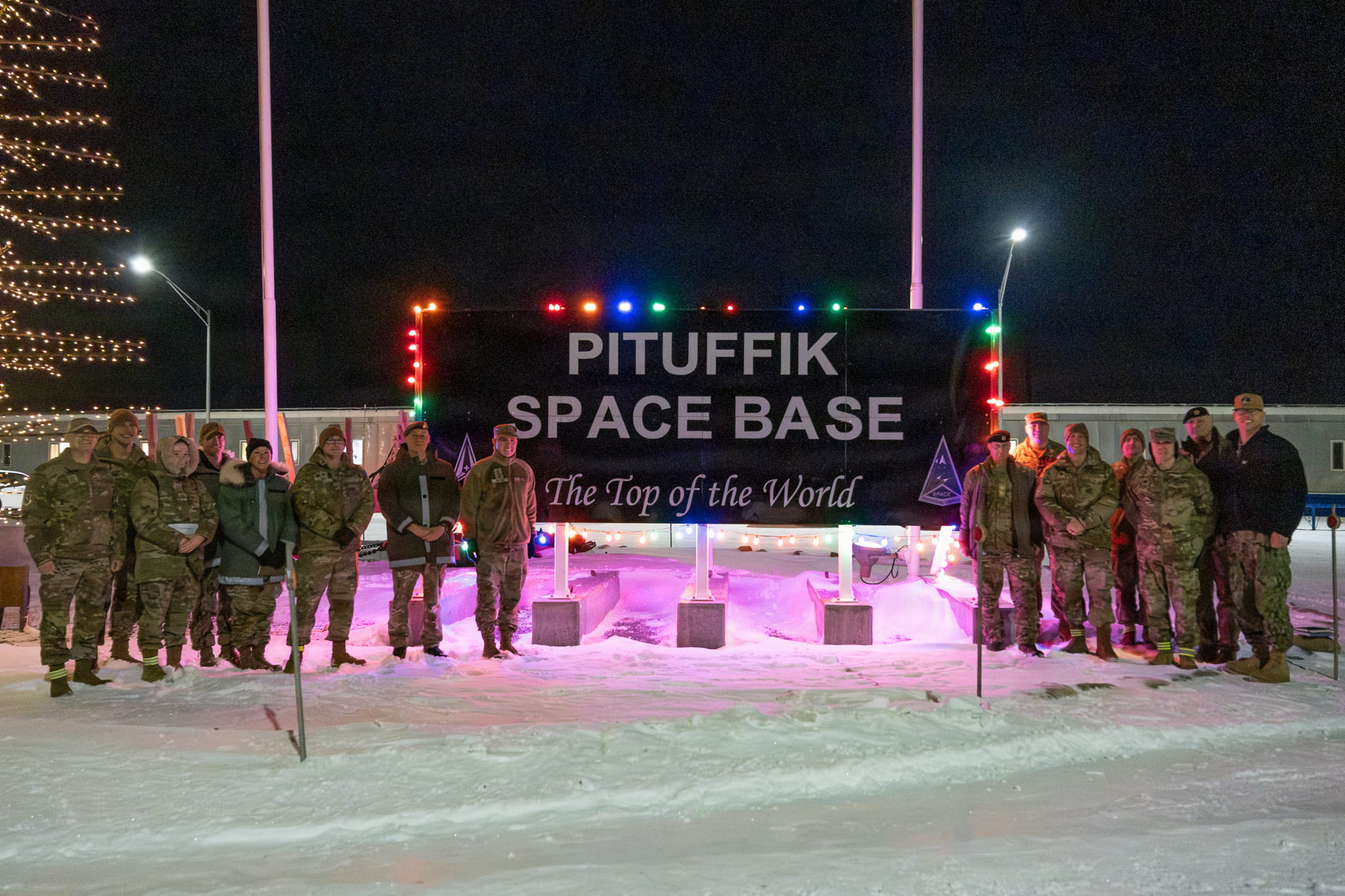 Group of military personnel posing in snowy conditions at night in front of the illuminated Pituffik Space Base sign in Greenland