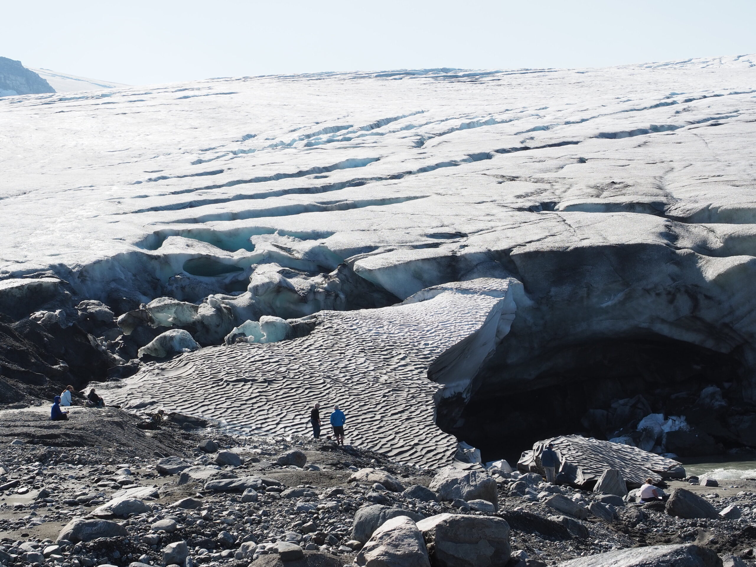 Snow-covered glacier with deep crevasses and an ice cave, with hikers standing on the rocky foreground