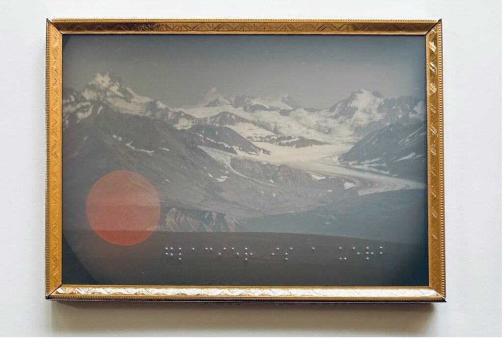 Framed vintage photograph of a snow-covered mountain range and glacier, with braille dots visible across the lower portion of the image