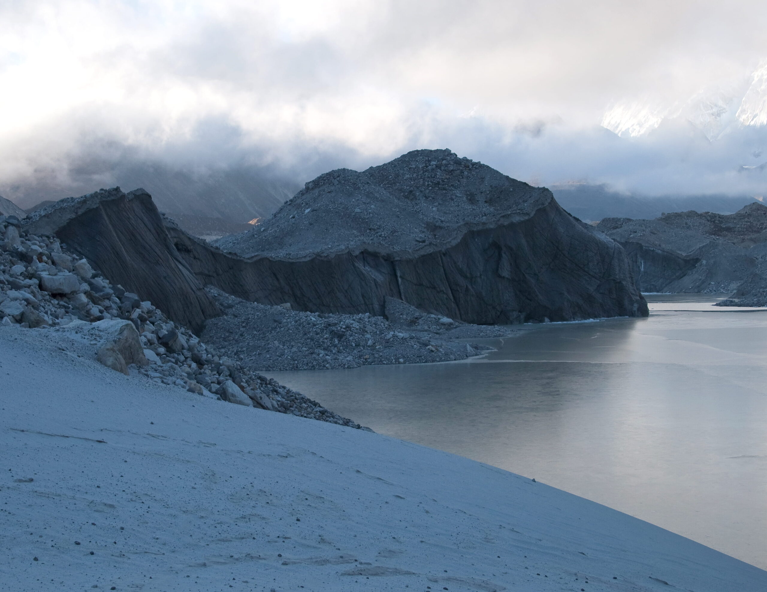 A frozen river runs between rugged granite peaks against an overcast sky