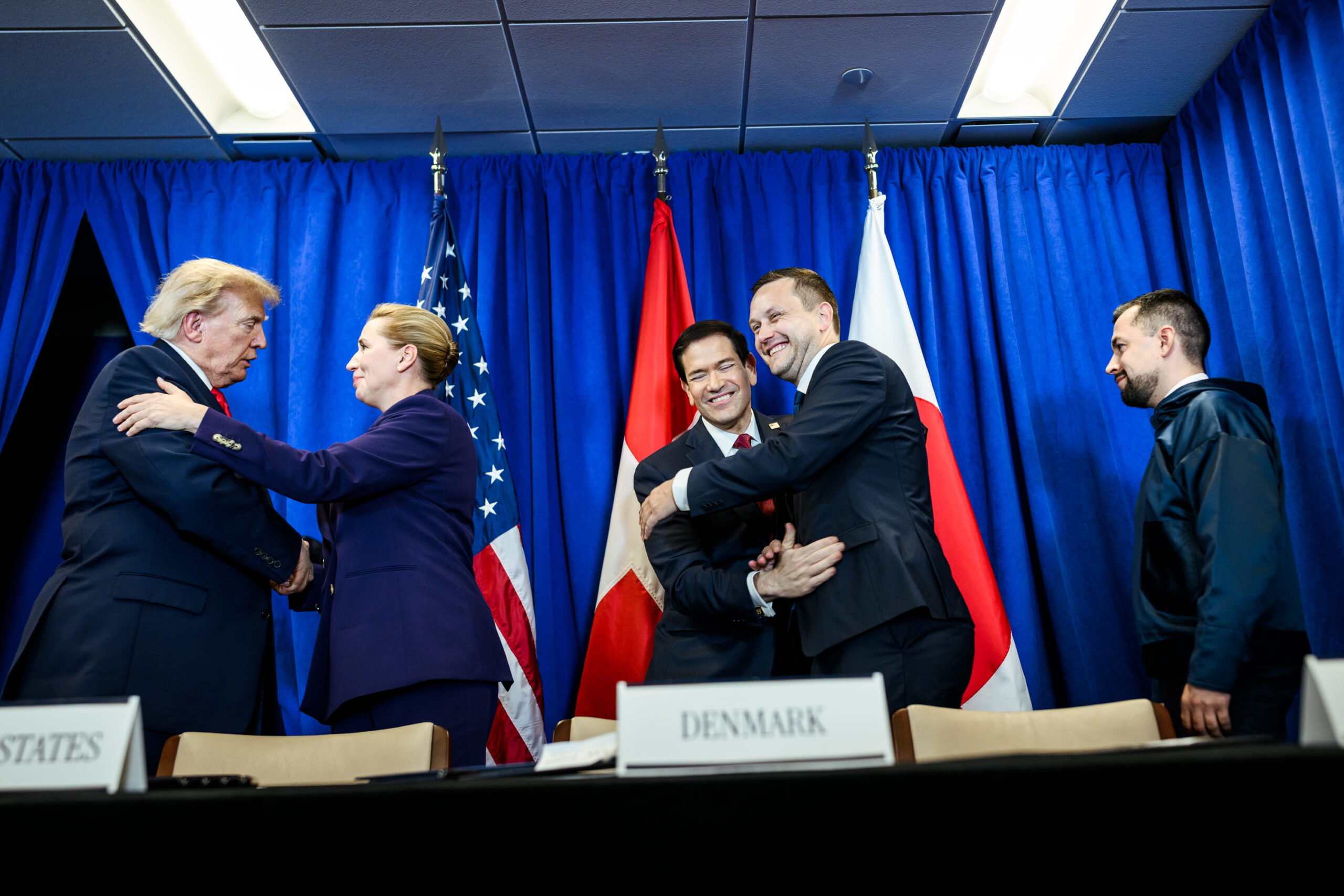 Donald Trump, Mette Frederiksen and Jan-Frederik Nielsen greet each other at a diplomatic meeting, with US, Danish, and Greenland flags in the background