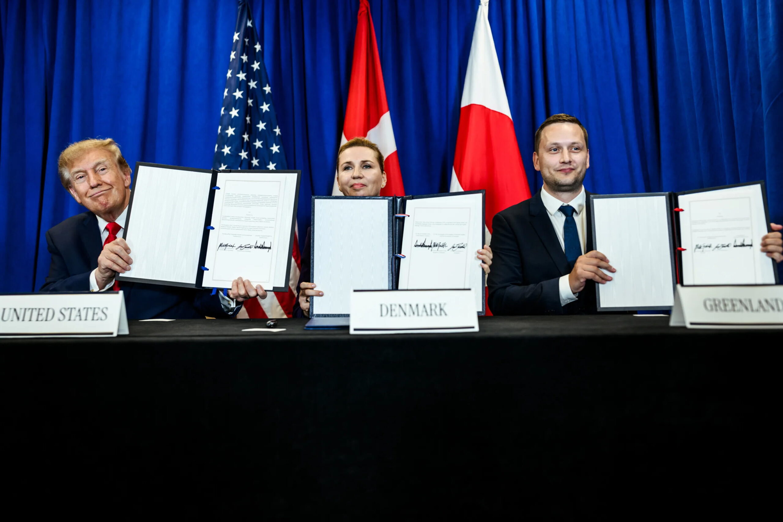 Donald Trump, Mette Frederiksen, and Jens-Frederik Nielsen hold signed documents at a diplomatic signing ceremony, seated in front of the United States, Denmark, and Greenland nameplates with national flags and blue curtains in the background
